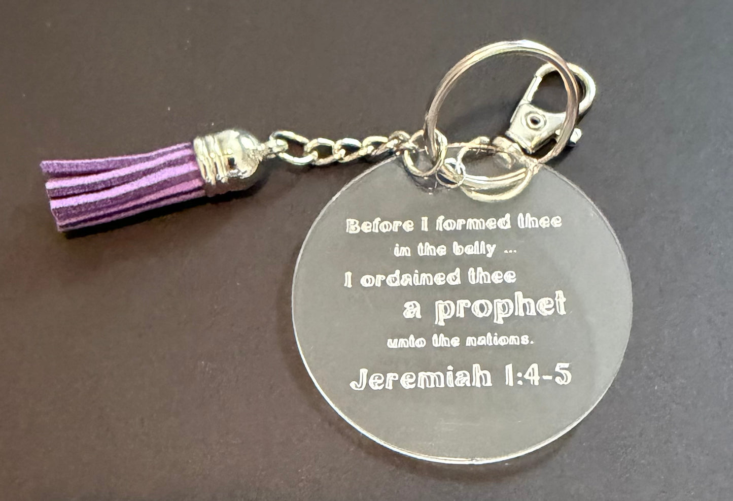 A round silver metal keychain with an engraved Bible verse from Isaiah 5:20, featuring a purple tassel charm, set against a dark background.