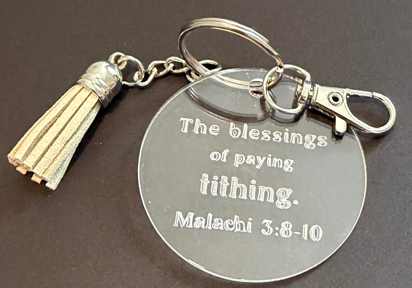 A round silver metal keychain with an engraved Bible verse from Isaiah 5:20, featuring a cream tassel charm, set against a dark background.