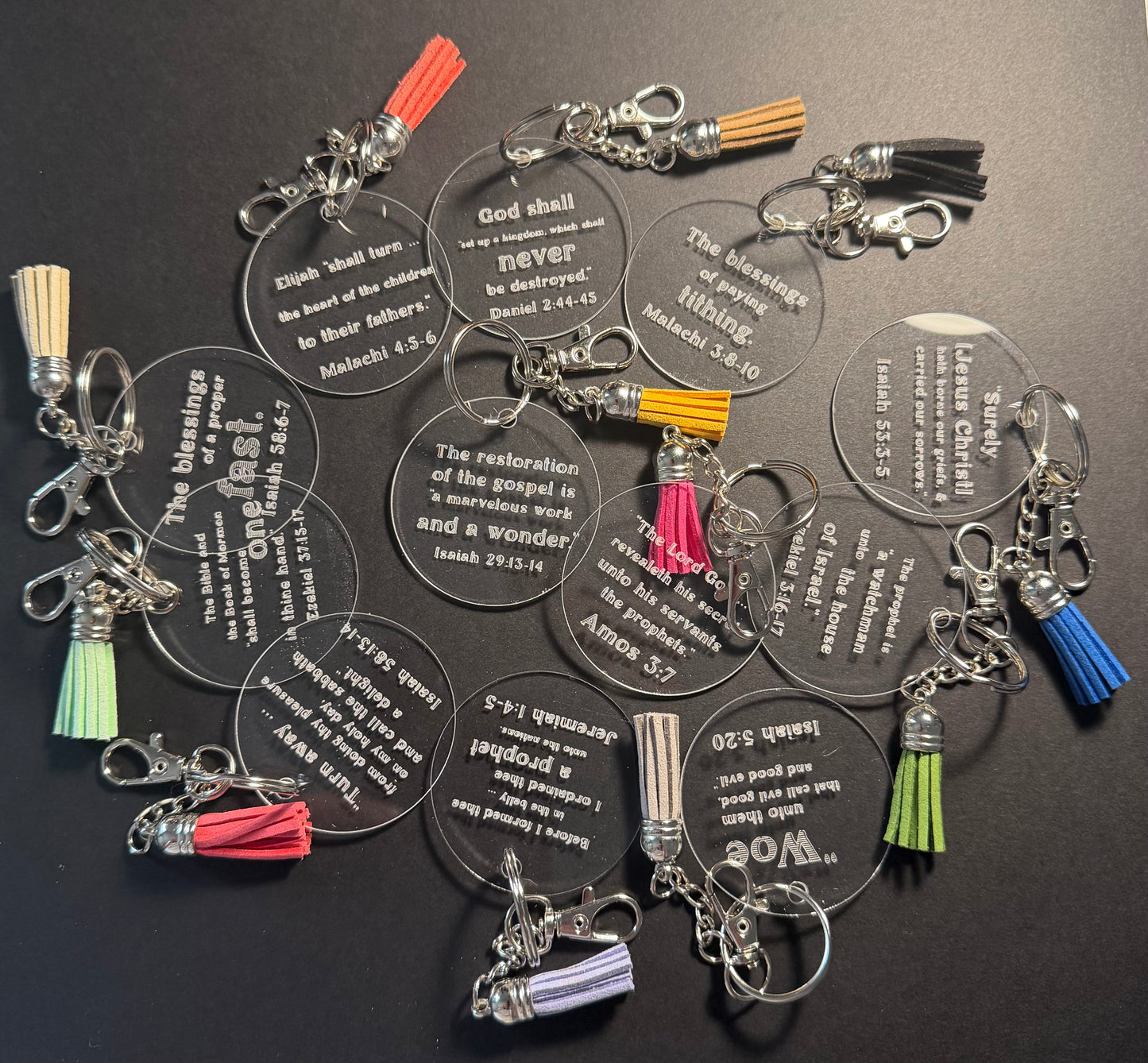A group of clear acrylic keychains featuring Bible verses, each with a silver lobster clasp and a colorful tassel in shades of red, blue, green, yellow, and purple, arranged on a dark background.