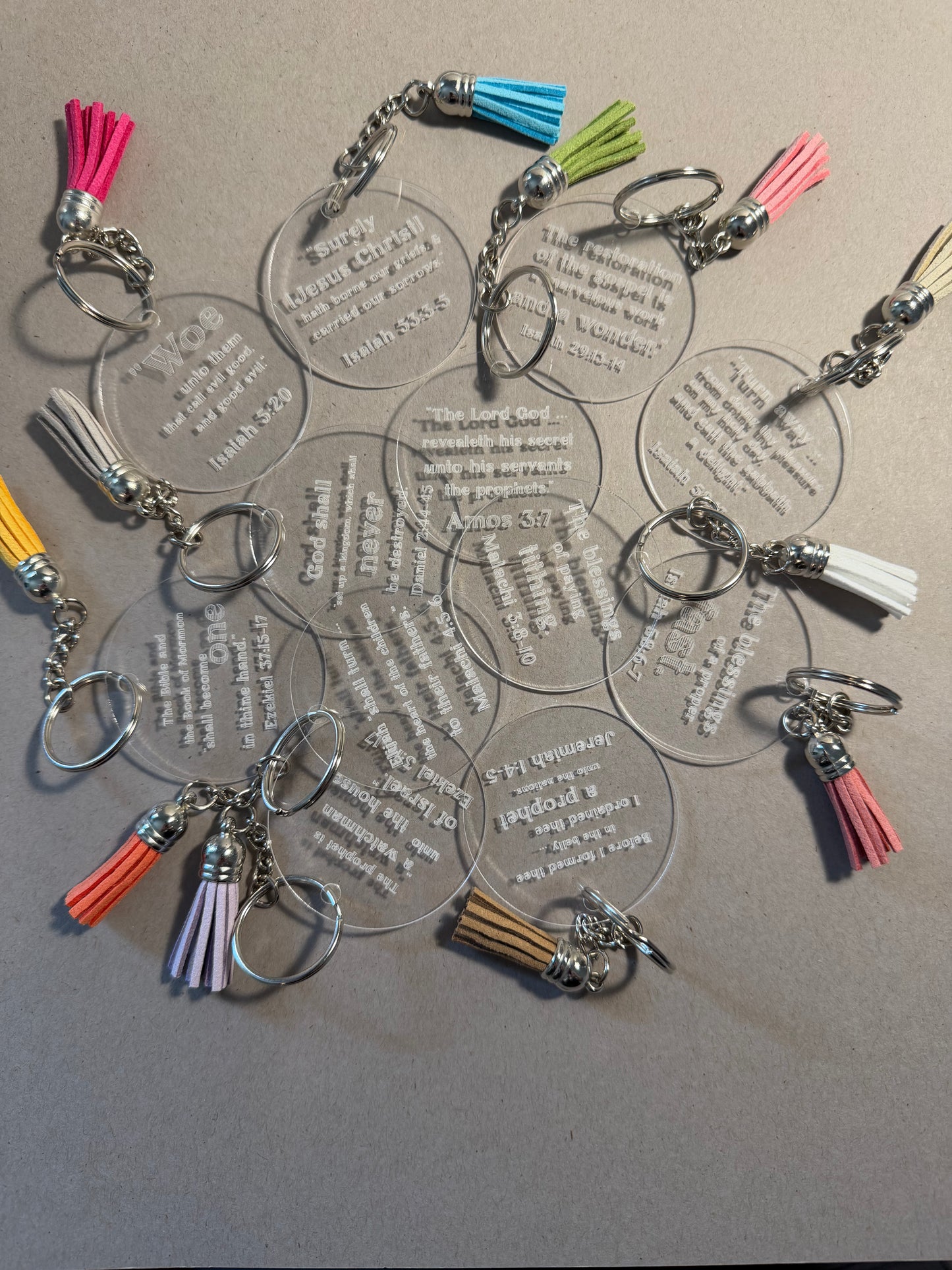 A group of clear acrylic keychains with silver metal rings and colorful tassels, arranged on a neutral gray background.