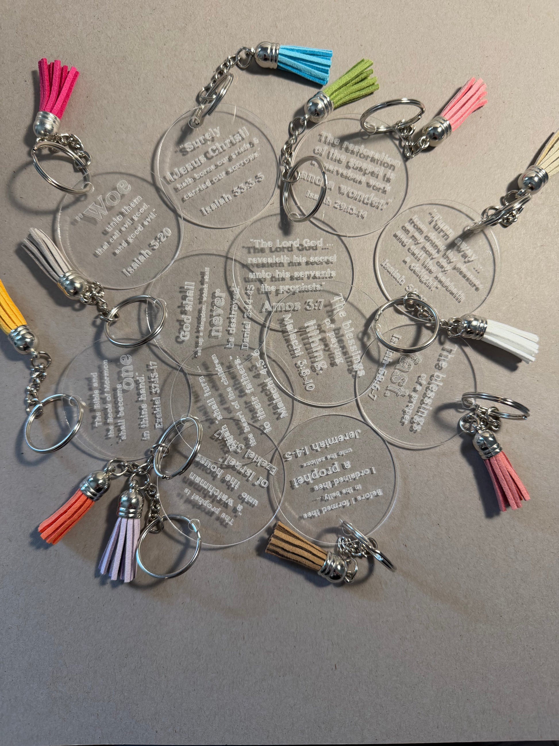 A group of clear acrylic keychains with silver metal rings and colorful tassels, arranged on a neutral gray background.