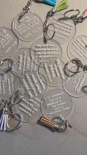 Several clear acrylic keychains with engraved Bible verses and silver key rings are arranged on a gray surface. Some keychains have colorful tassels in blue, green, pink, purple, and brown.