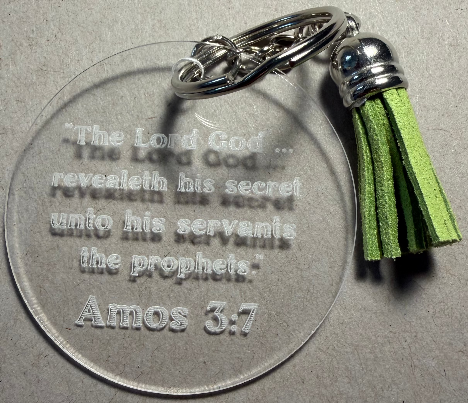 A clear acrylic keychain featuring an engraved Bible verse from Amos 3:7, attached to a silver split ring and a green tassel charm with a silver cap, set against a textured gray background.