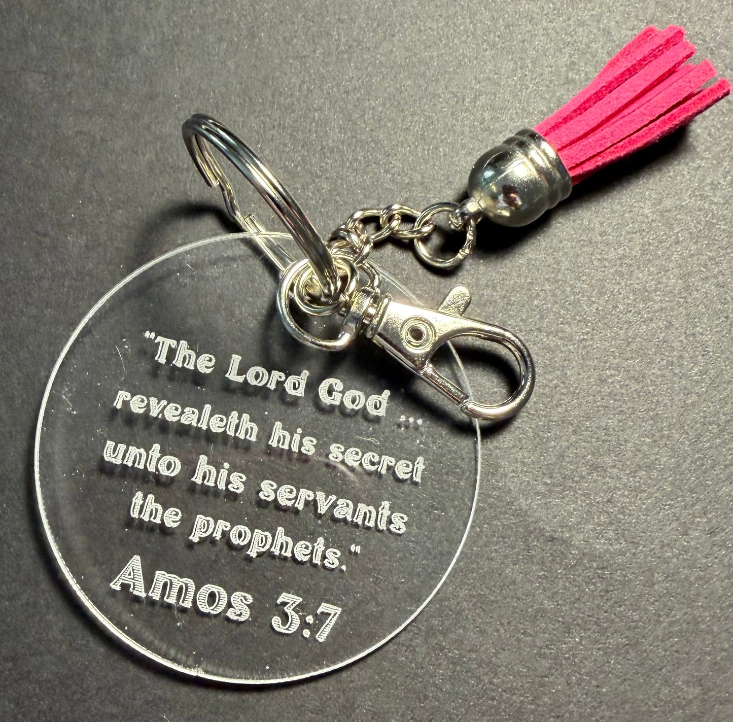 A round clear acrylic keychain featuring an engraved Bible verse from Amos 3:7, attached to a silver metal split ring, clasp, and chain, with a bright pink tassel on a dark gray textured background.