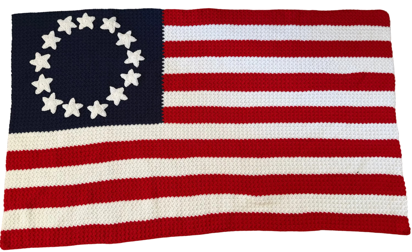 A crocheted American flag rug featuring 13 red and white stripes and a navy blue canton with 13 white stars arranged in a circle, laid flat on a patterned gray rug.