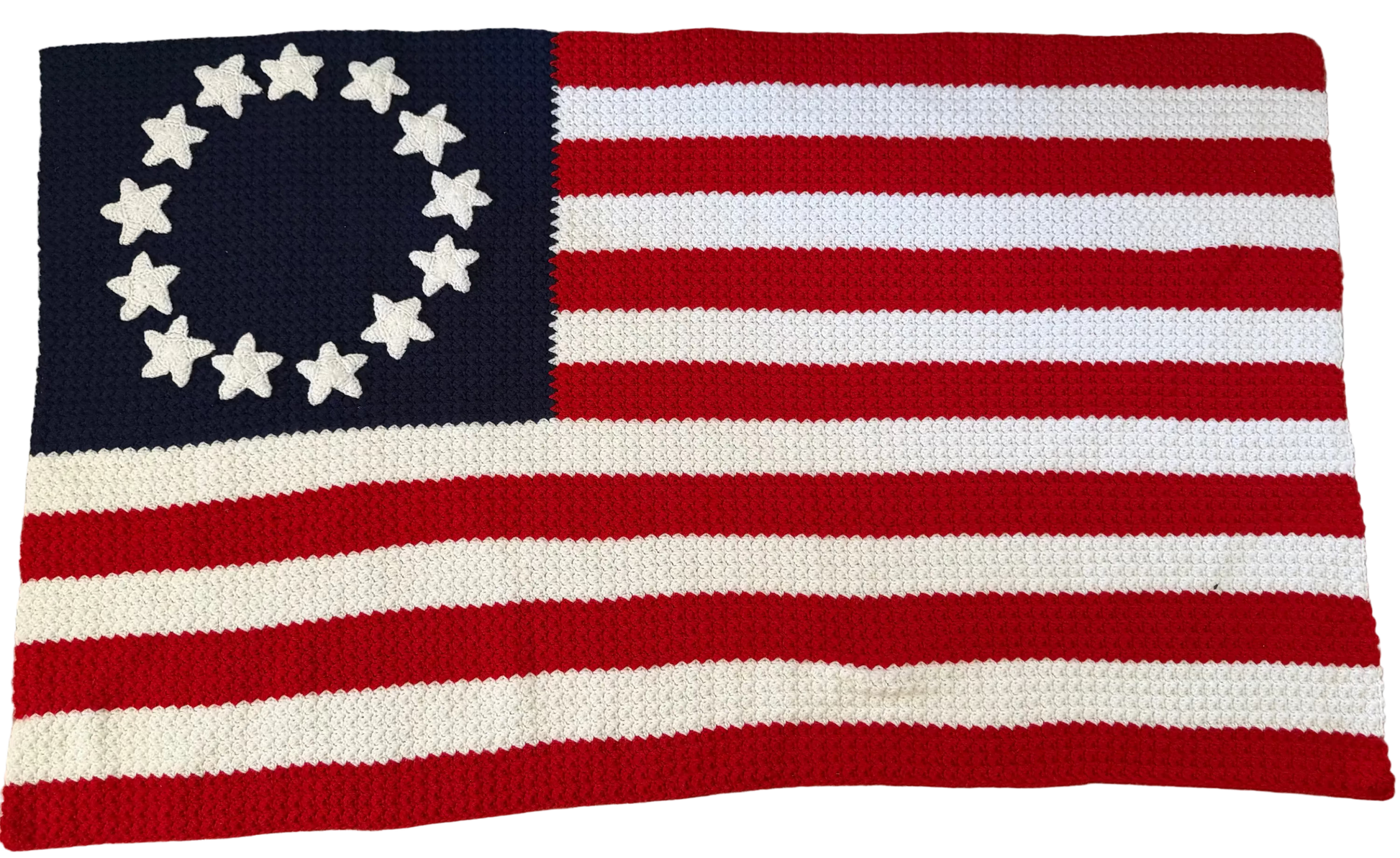 A crocheted American flag rug featuring 13 red and white stripes and a navy blue canton with 13 white stars arranged in a circle, laid flat on a patterned gray rug.