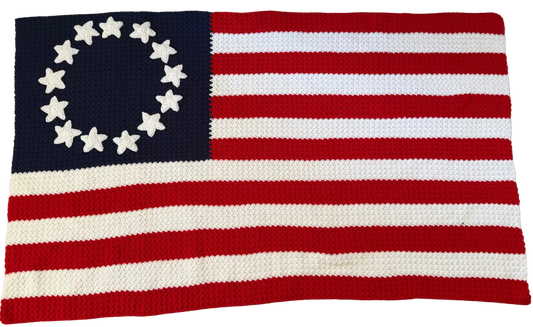 A crocheted American flag rug featuring 13 red and white stripes and a navy blue canton with 13 white stars arranged in a circle, laid flat on a patterned gray rug.