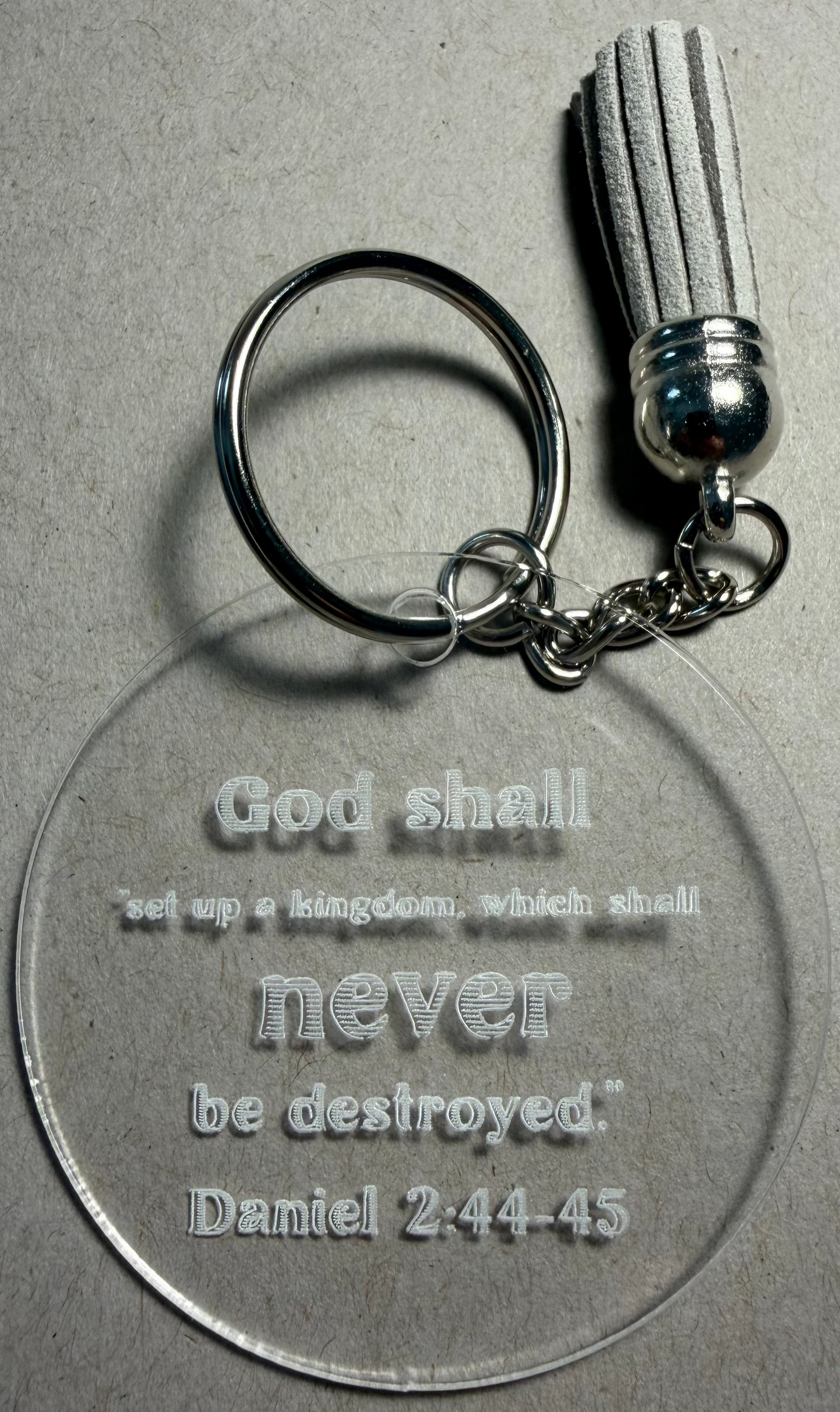 A clear acrylic keychain with a silver split ring and tassel, featuring the engraved text 'God shall set up a kingdom, which shall never be destroyed. Daniel 2:44-45' on a textured gray background.