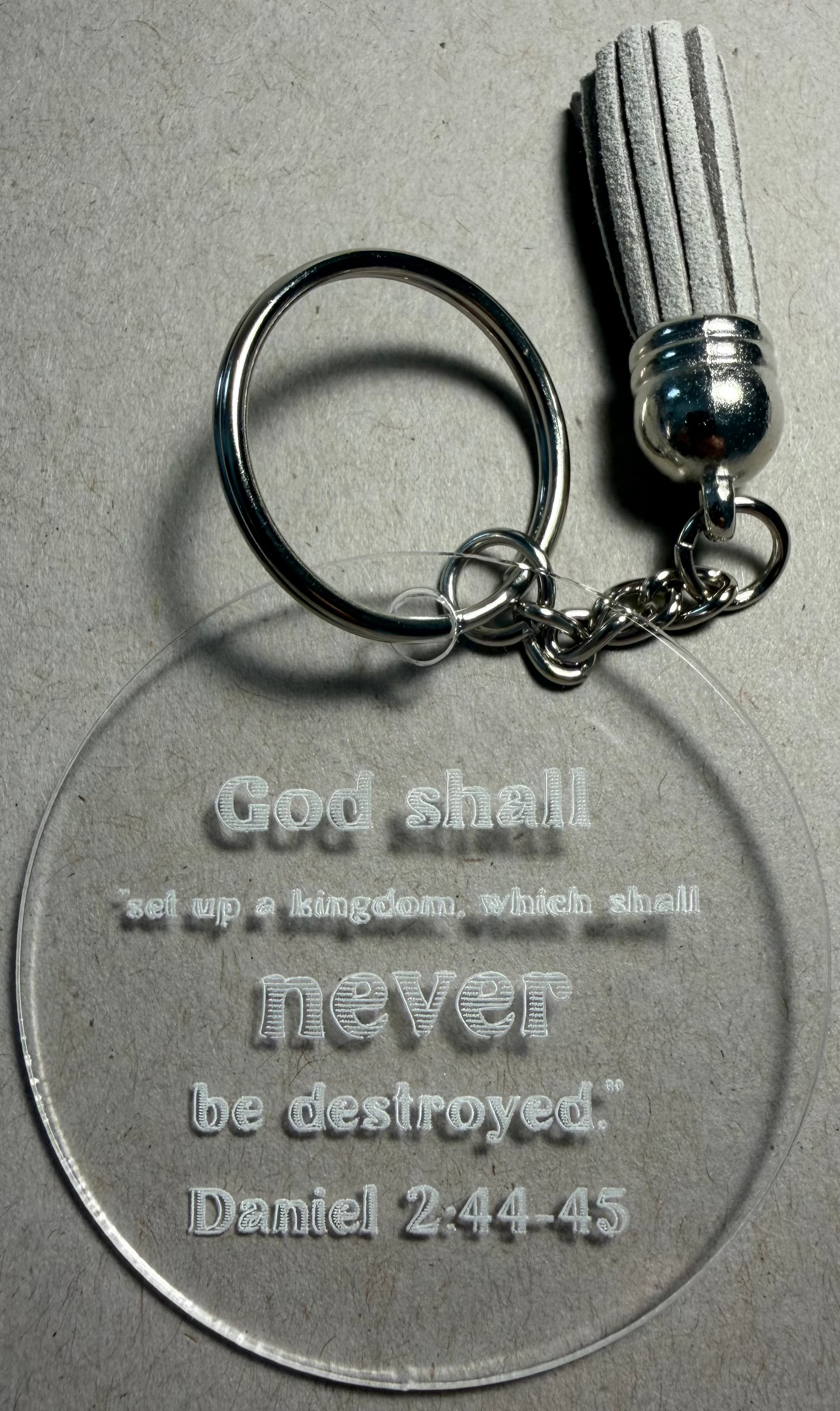 A clear acrylic keychain with a silver split ring and tassel, featuring the engraved text 'God shall set up a kingdom, which shall never be destroyed. Daniel 2:44-45' on a textured gray background.