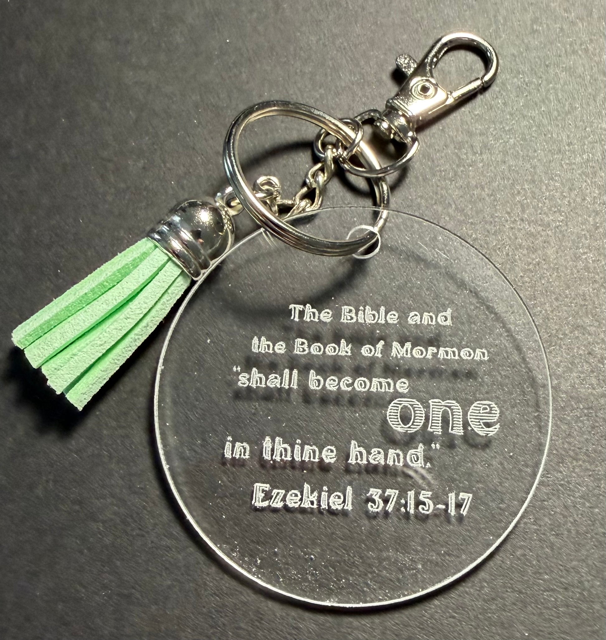 A clear acrylic keychain with a silver-tone metal clasp, key ring, and a light green tassel. The round clear tag is engraved with the Bible verse, 'The Bible and the Book of Mormon shall become one in thine hand.