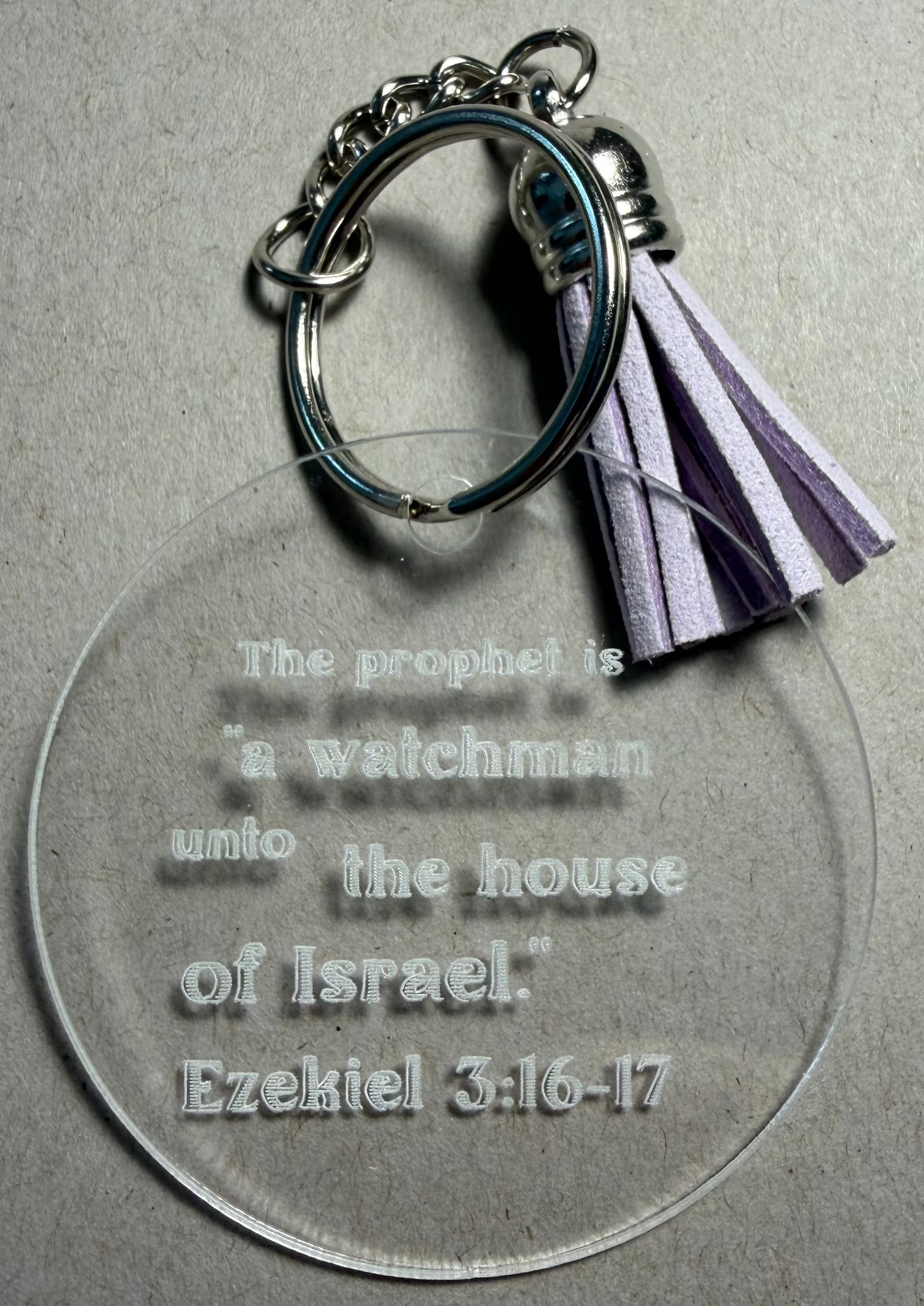 A clear acrylic keychain with an engraved Bible verse from Ezekiel 3:16-17, featuring a silver metal key ring and a purple and white striped tassel, shown on a textured gray background.