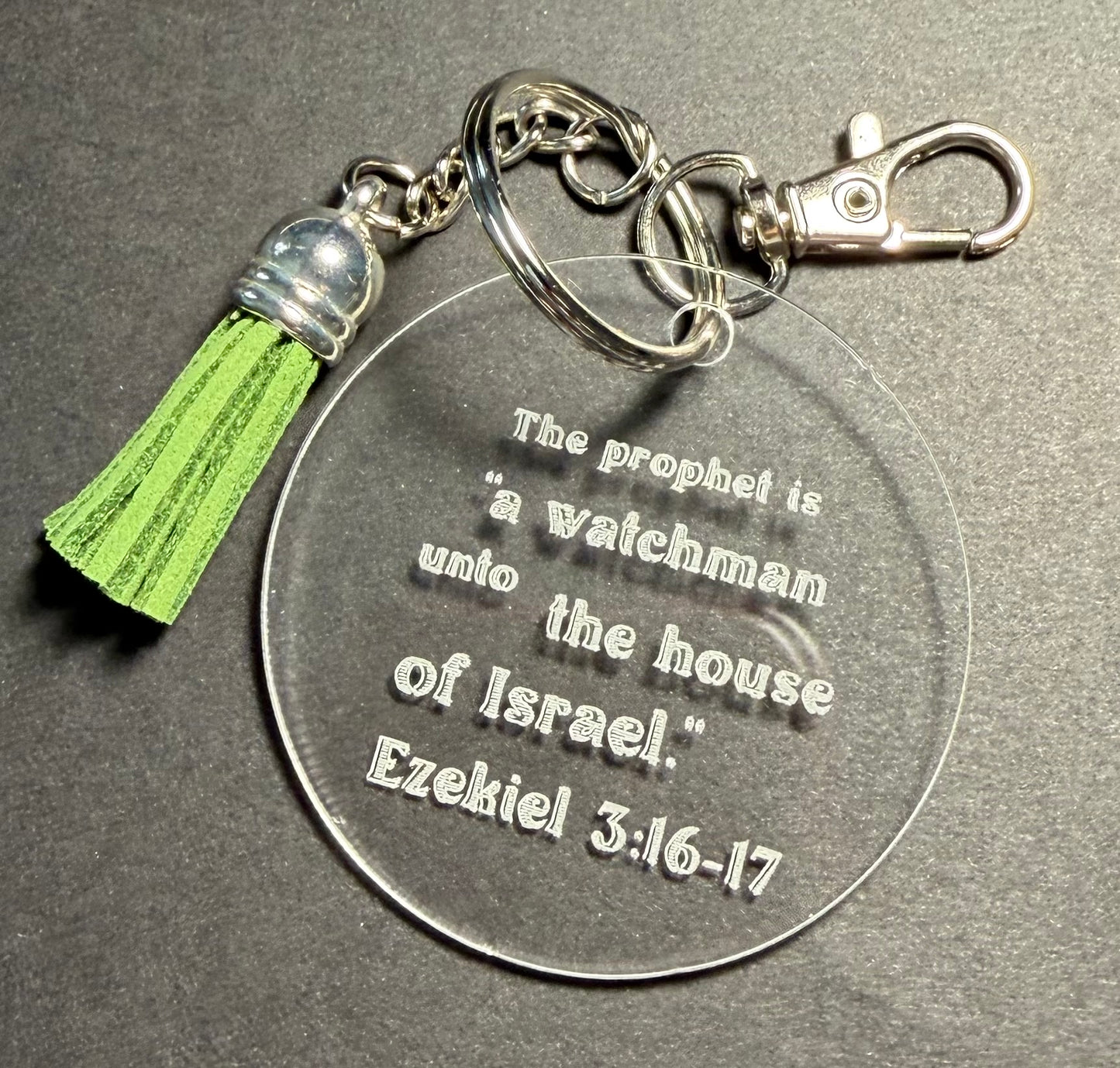A clear acrylic keychain with engraved text reading 'The prophet is "a watchman unto the house of Israel." Ezekiel 3:16-17,' attached to a silver-tone split ring, lobster clasp, and a green tassel with a silver bead cap, shown on a dark gray textured background.