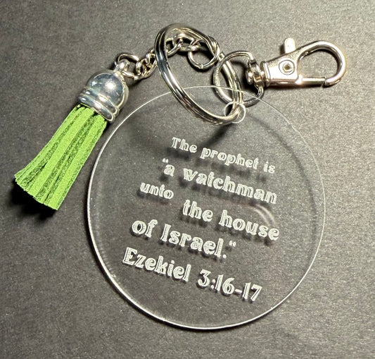 A clear acrylic keychain with engraved text reading 'The prophet is "a watchman unto the house of Israel." Ezekiel 3:16-17,' attached to a silver-tone split ring, lobster clasp, and a green tassel with a silver bead cap, shown on a dark gray textured background.
