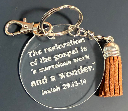 A round clear acrylic keychain with an engraved Bible verse, attached to a silver-tone split ring and lobster clasp, with a brown suede tassel charm on a dark background.