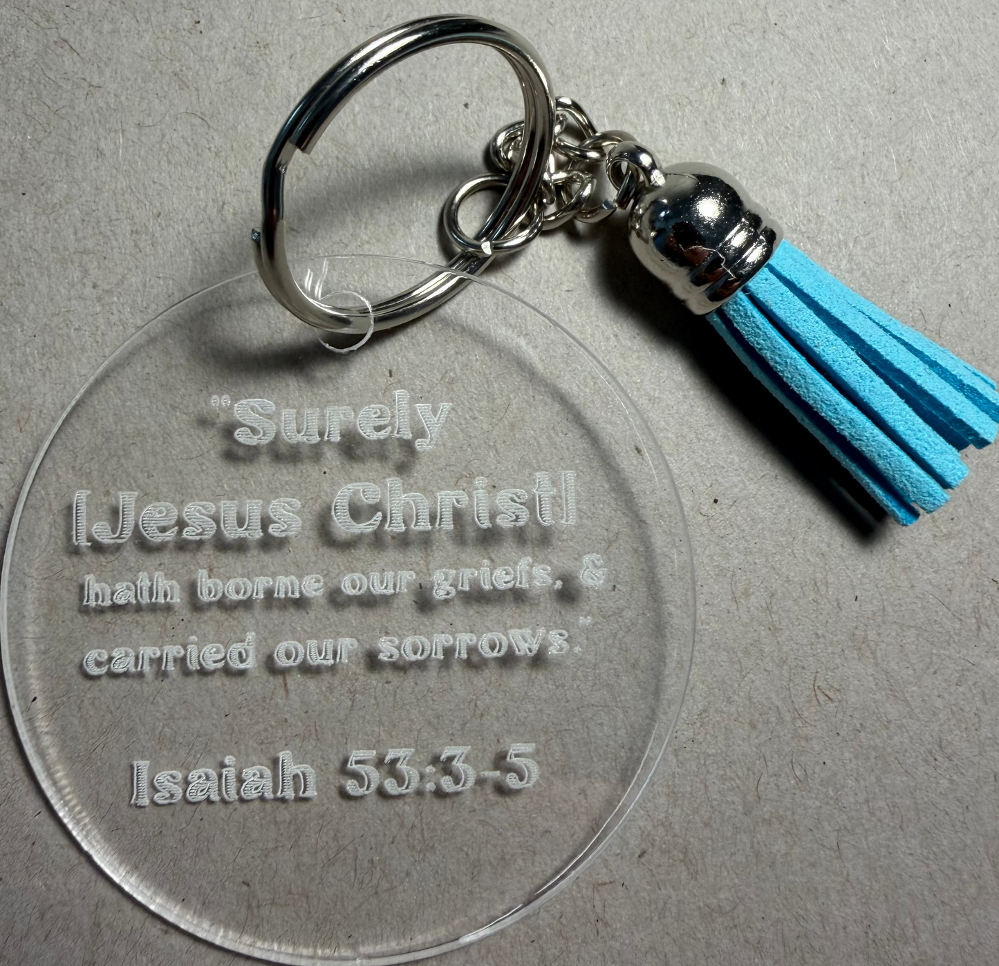 A clear acrylic keychain with a silver metal split ring and a bright blue tassel charm, engraved with the Bible verse Isaiah 53:3-5.