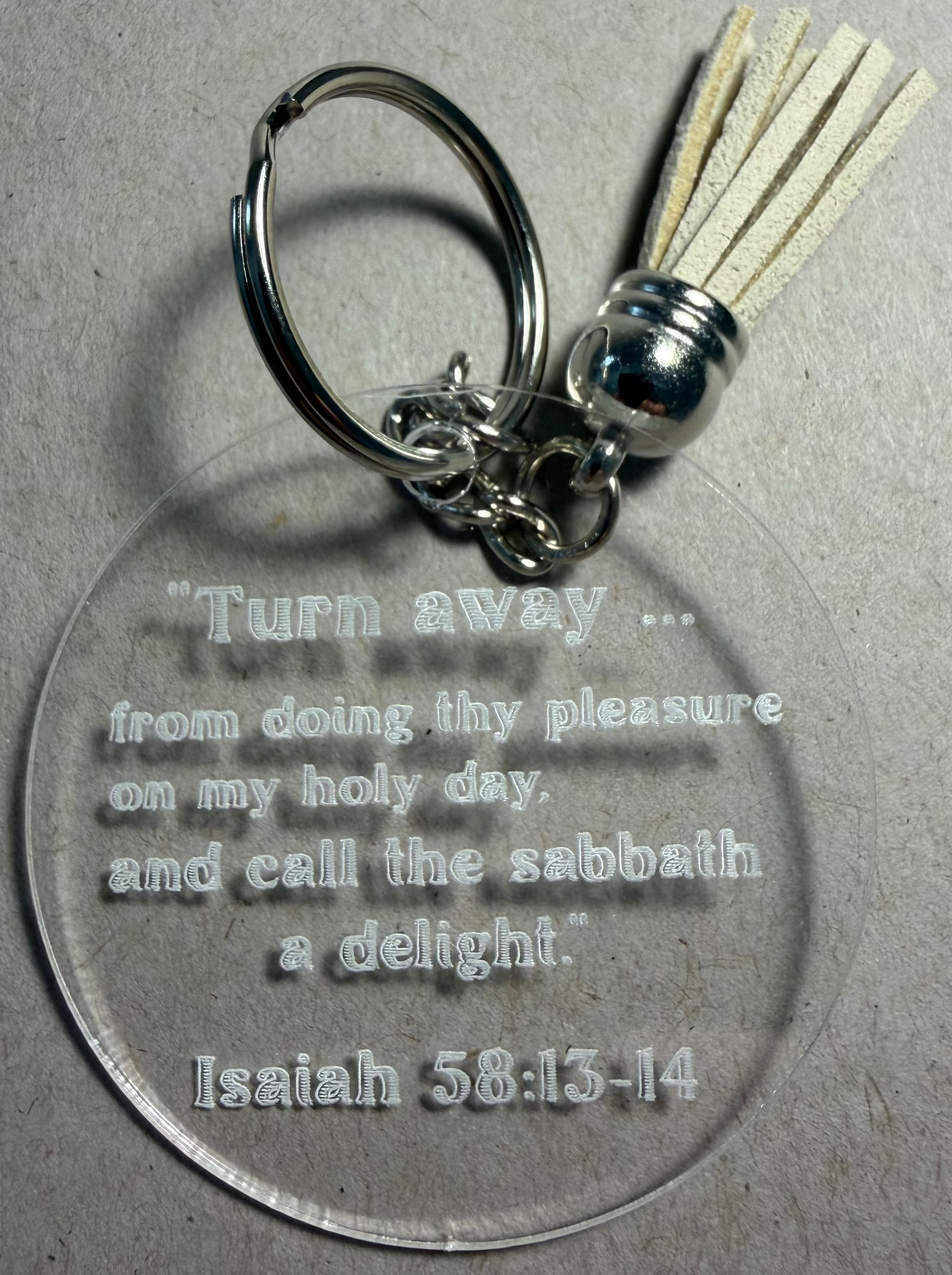 A clear acrylic keychain with a silver metal split ring and tassel, featuring an engraved biblical quote from Isaiah 58:13-14 on a gray textured background.