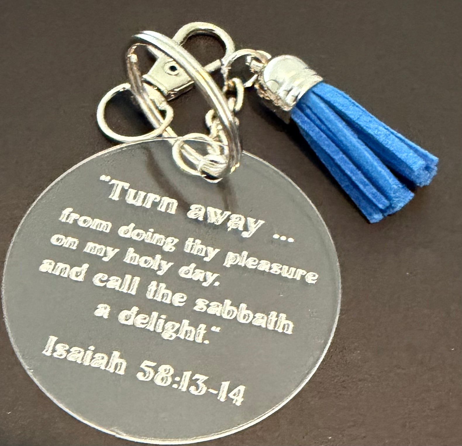 A round silver metal keychain with an engraved Bible verse from Isaiah 5:20, featuring a pink and white tassel charm, set against a dark background.