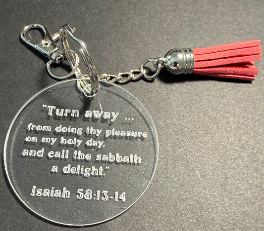 A round clear acrylic keychain featuring an engraved biblical quote from Isaiah 58:13-14, attached to a silver metal clasp and chain with a red tassel, shown on a dark gray textured background.