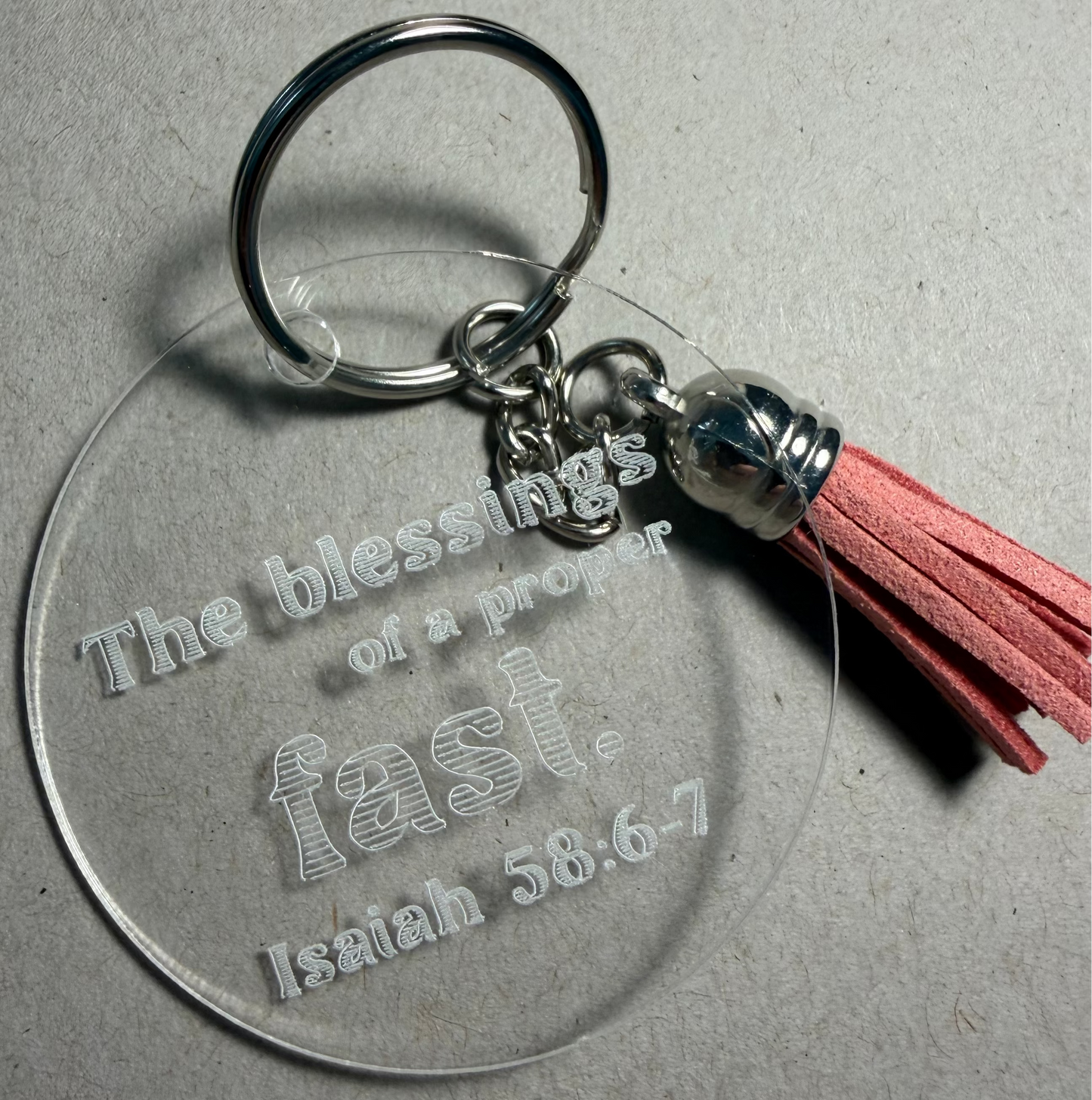 A clear acrylic keychain with an engraved biblical quote from Isaiah 58:13-14, attached to a silver metal split ring and a cream-colored tassel with a silver bead, set against a textured gray background.