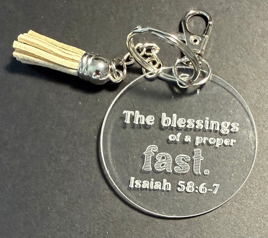 A clear acrylic round keychain with engraved text reading 'The blessings of a proper fast. Isaiah 58:6-7', attached to a silver split ring, lobster clasp, and a cream tassel on a dark gray background.