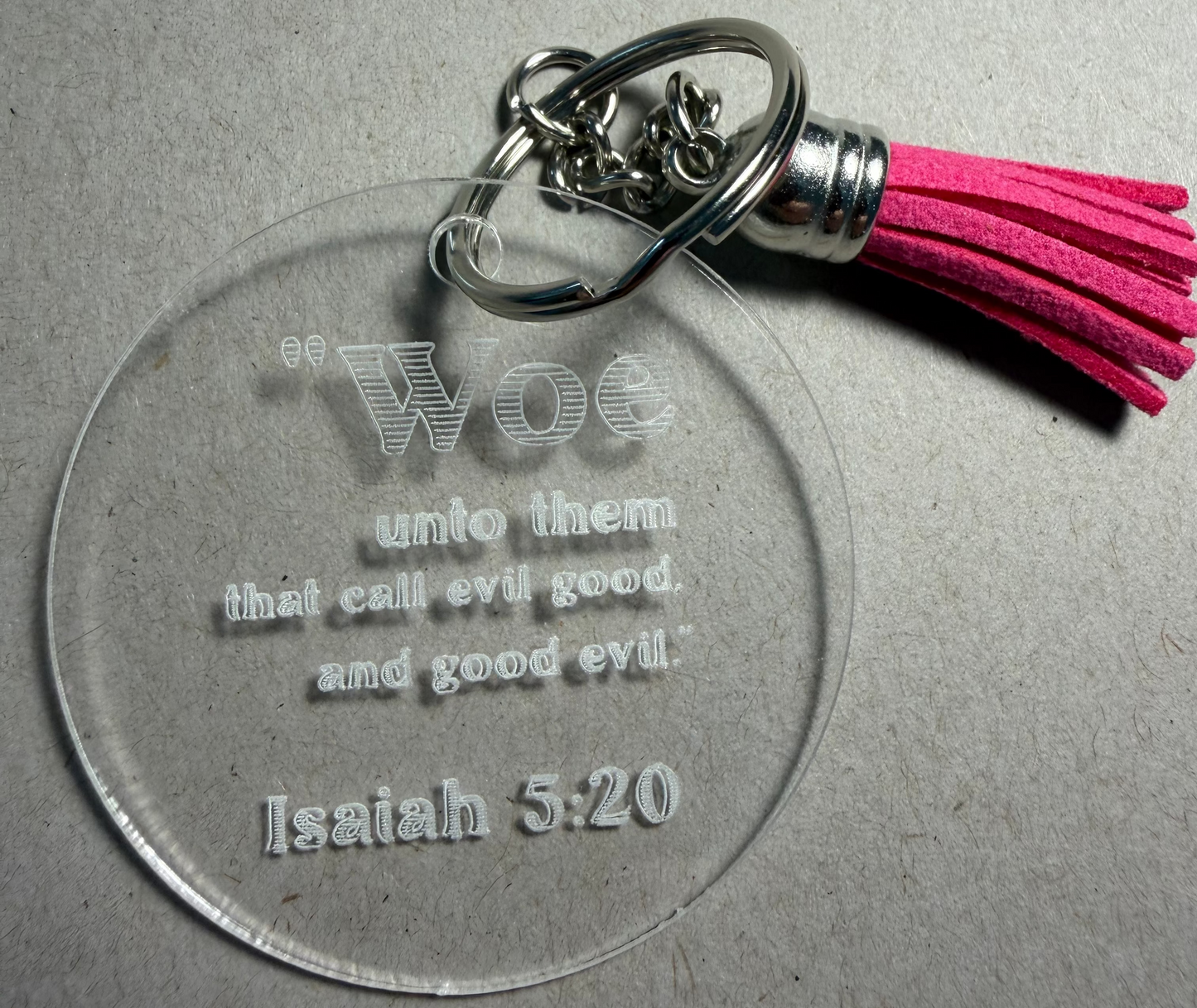 A clear acrylic keychain featuring the engraved text 'Woe unto them that call evil good, and good evil; Isaiah 5:20' with a bright pink tassel and silver metal keyring, shown on a textured gray background.