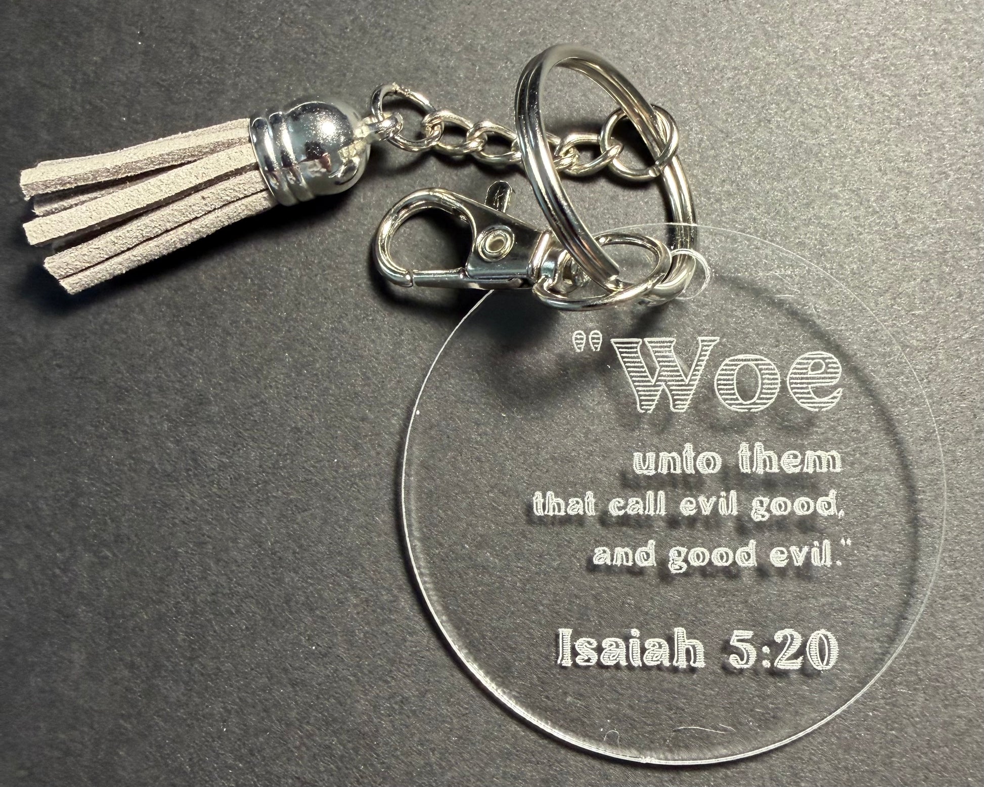A clear acrylic keychain with a silver-tone metal clasp, split ring, and tassel. The round clear disc is engraved with the quote: 'Woe unto them that call evil good, and good evil.