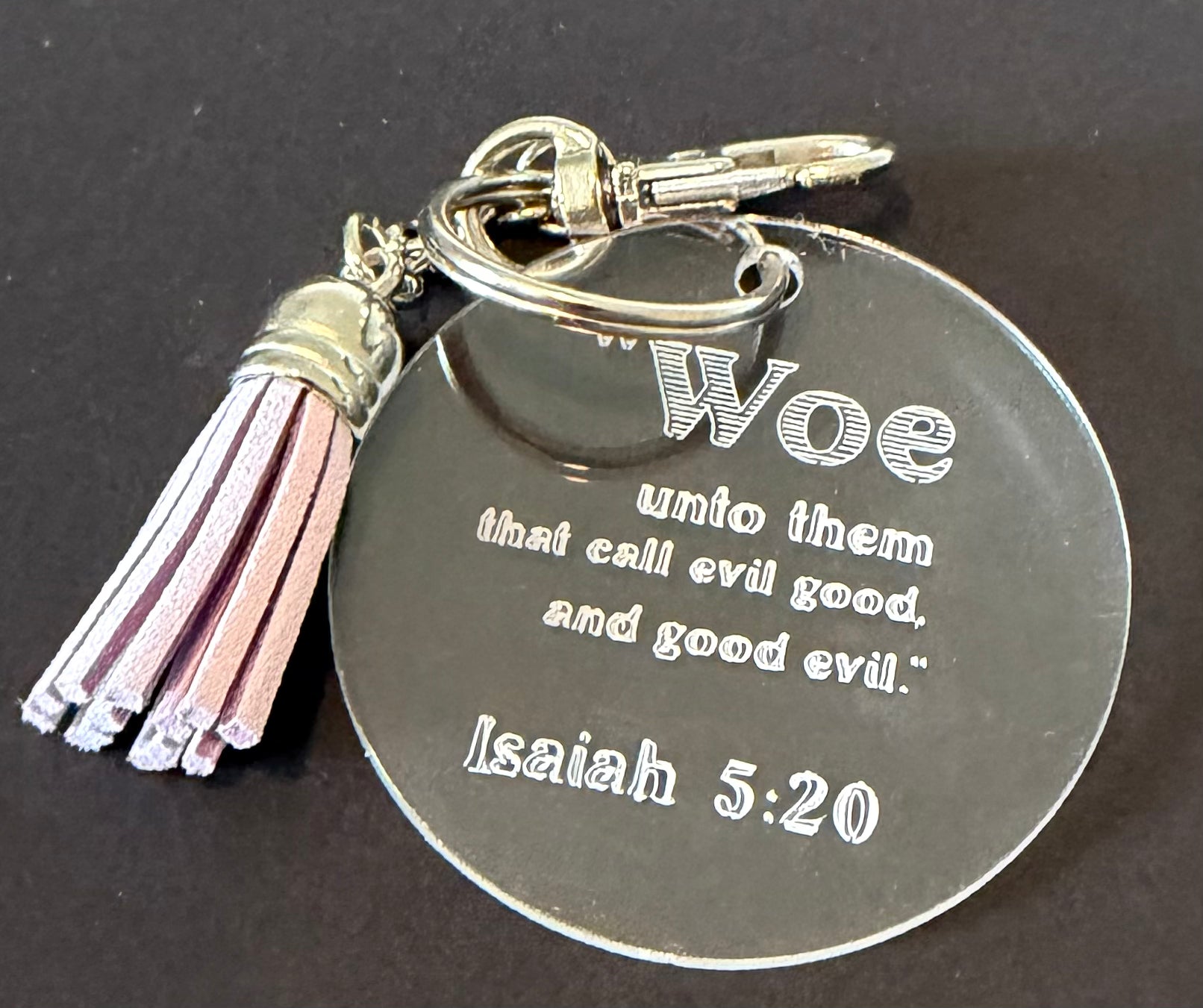 A round silver metal keychain with an engraved Bible verse from Isaiah 5:20, featuring a pink and white tassel charm, set against a dark background.