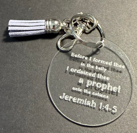 A clear acrylic round keychain with engraved text from Jeremiah 1:4-5, attached to silver metal rings and a lilac tassel, shown on a dark gray background.