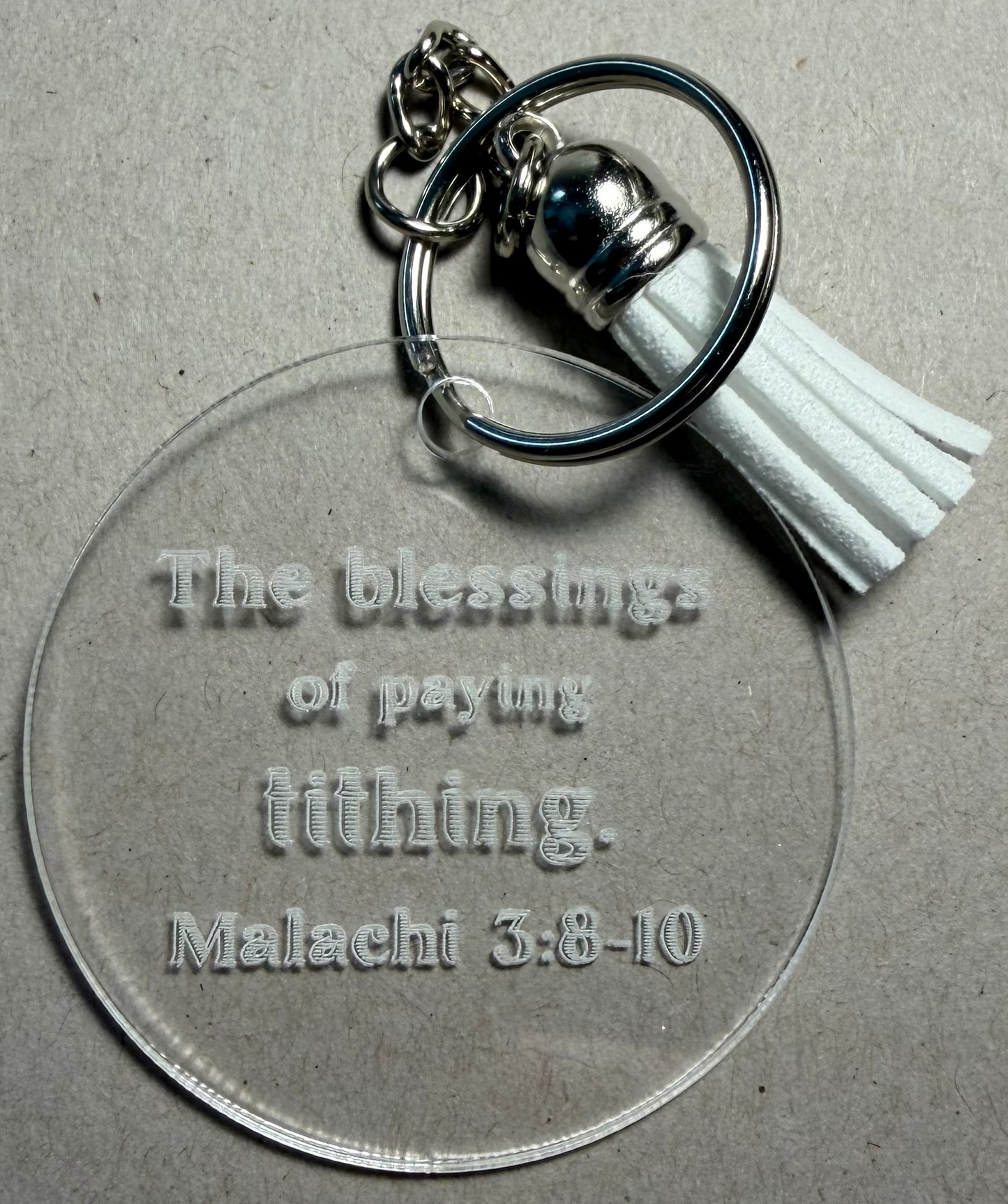 A clear acrylic keychain featuring an engraved Bible verse from Malachi 3:8-10, complete with a silver metal key ring and a white tassel, set against a textured gray background.