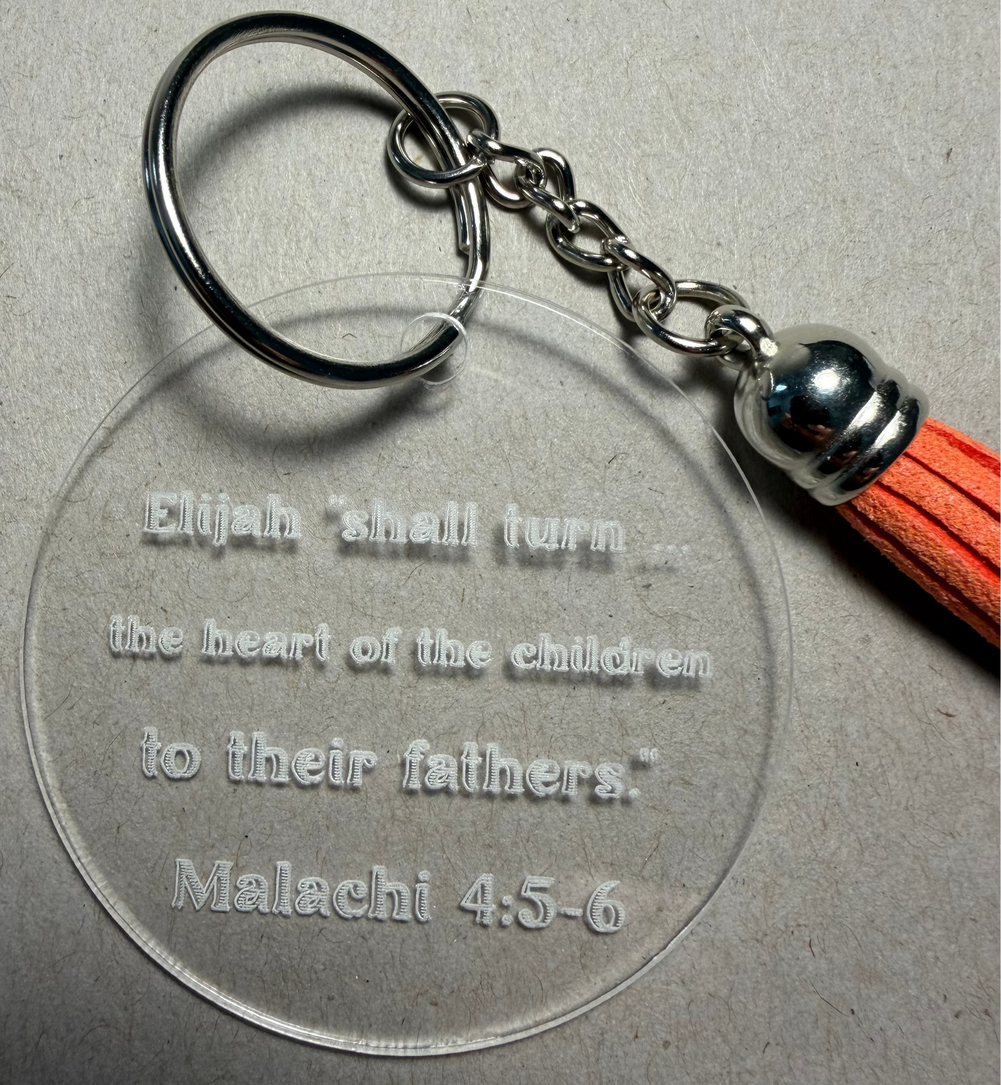 A clear acrylic keychain with an engraved Bible verse, attached to a silver metal split ring and chain with an orange tassel, shown on a neutral gray background.