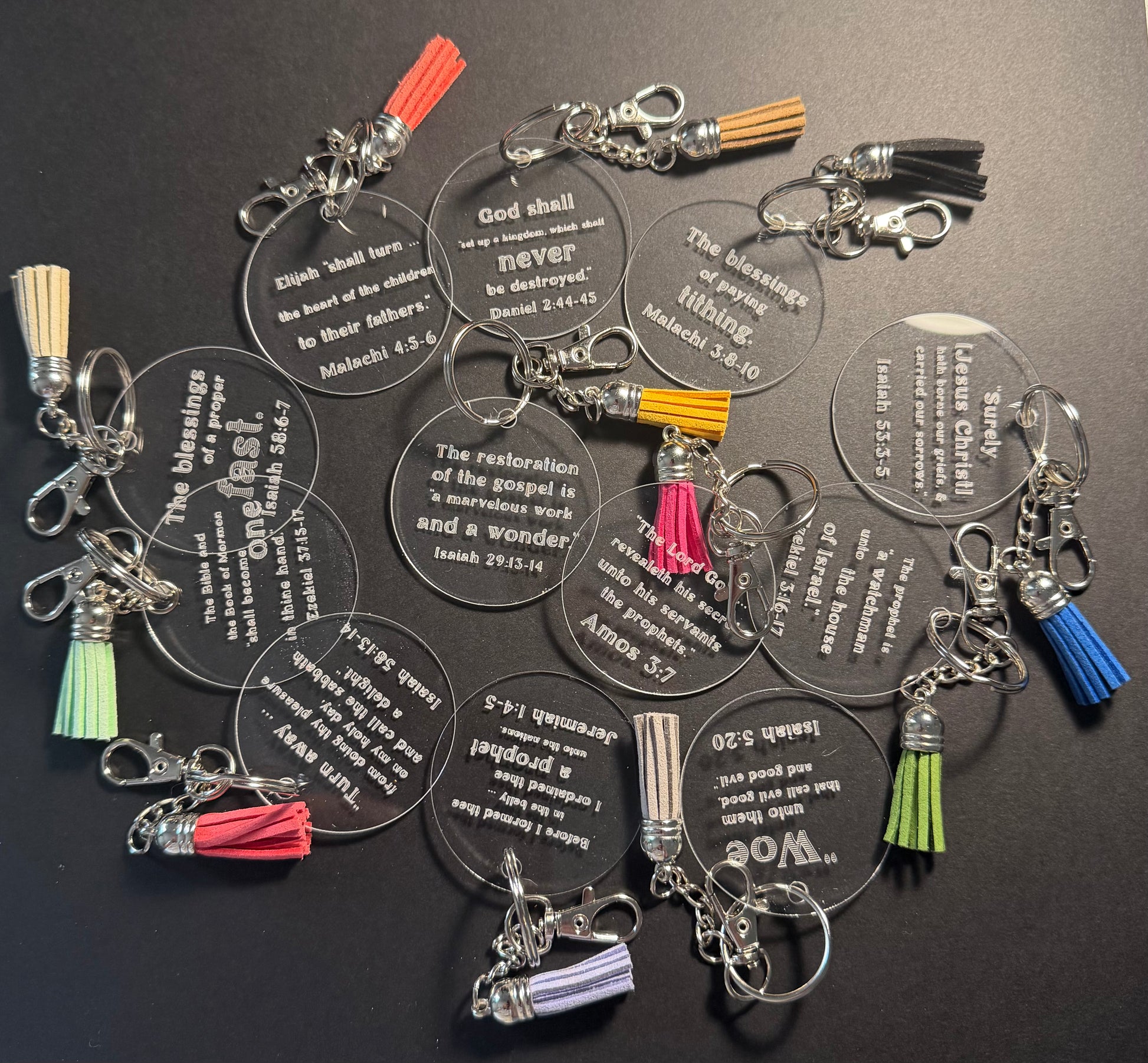A group of clear acrylic keychains featuring Bible verses, each with a silver lobster clasp and a colorful tassel in shades of red, blue, green, yellow, and purple, arranged on a dark background.